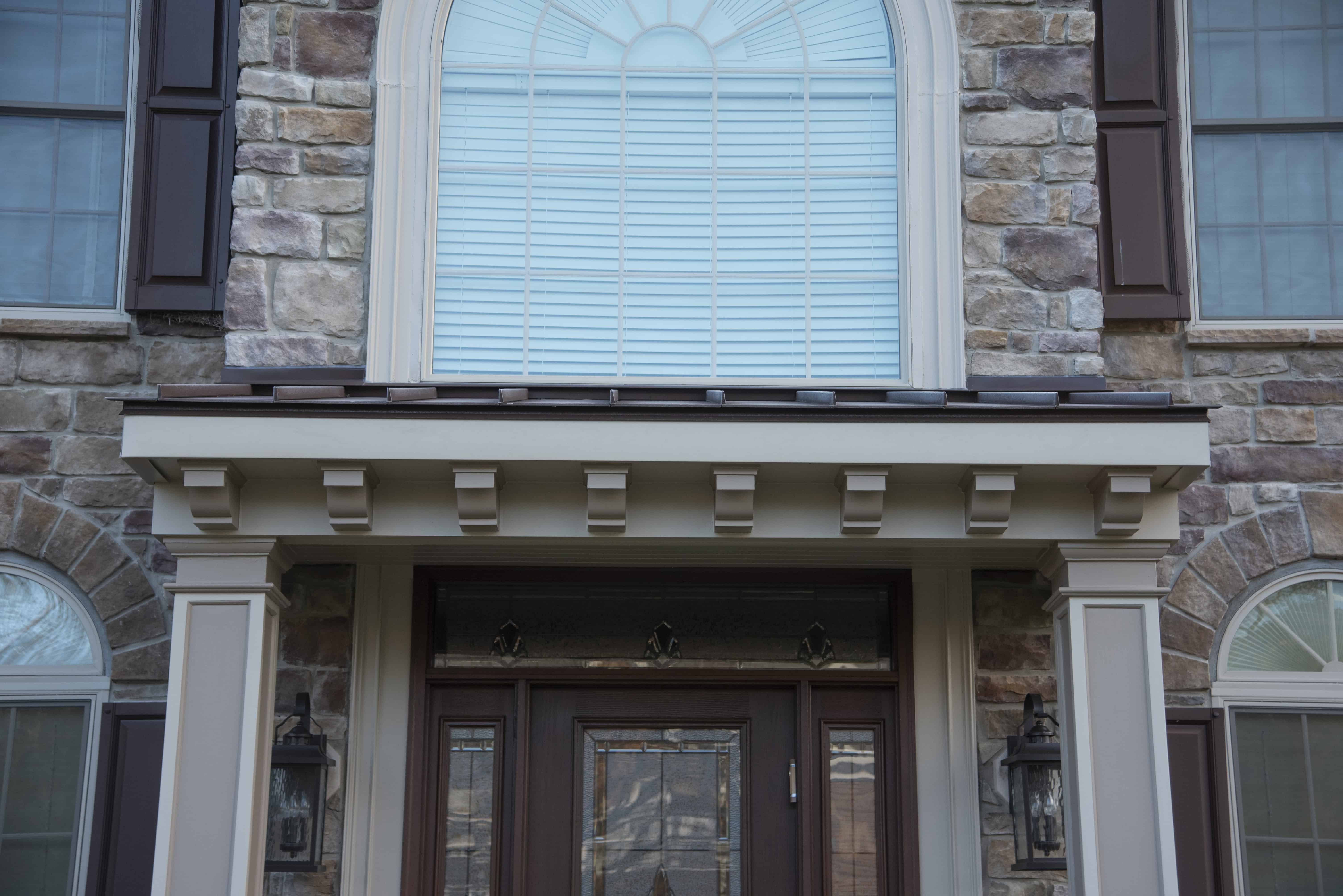 Close-up of a house entrance featuring a stone exterior, brown double doors with decorative glass, two wall lanterns, and a large arched window with white blinds above the doorway.