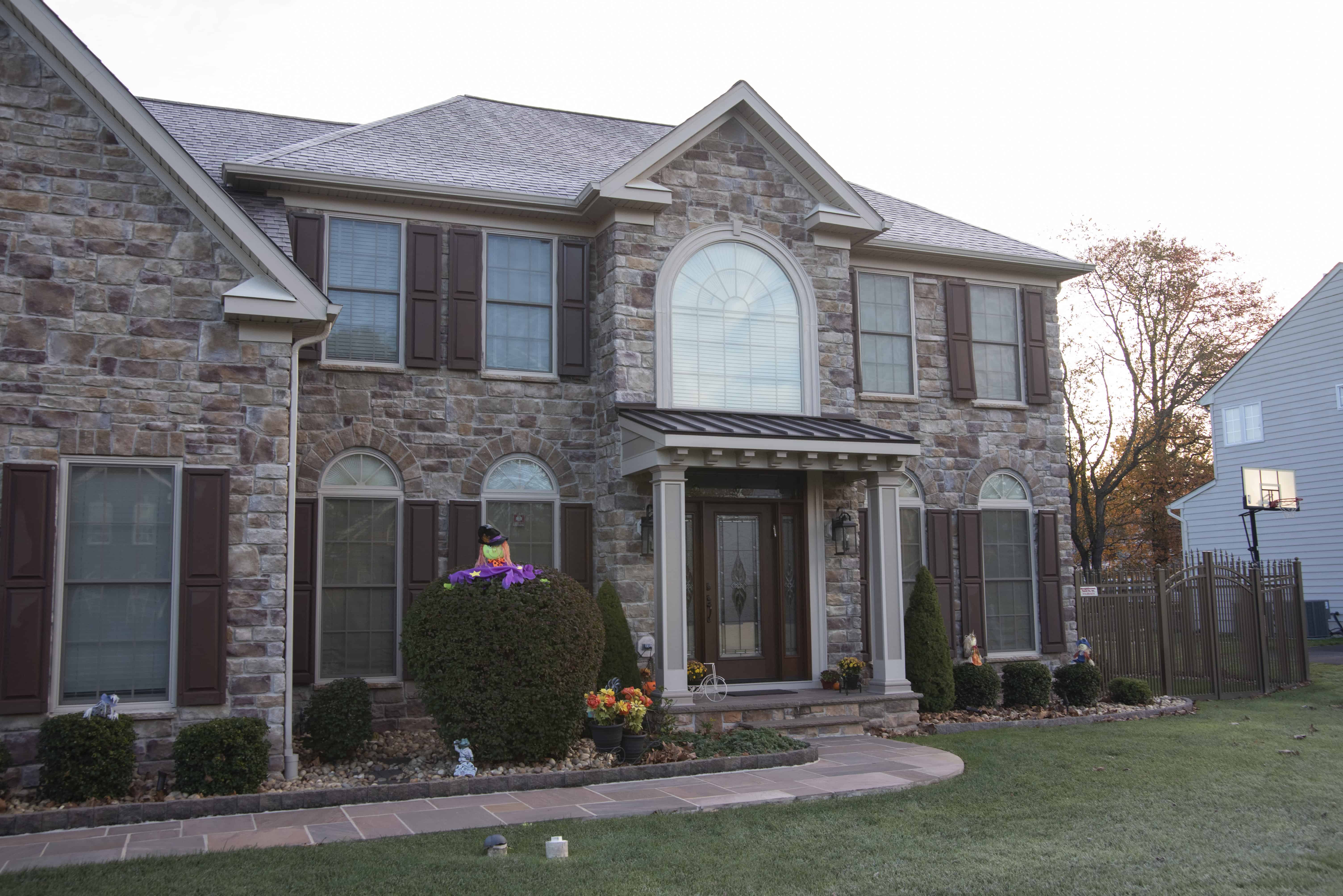 A two-story stone house with brown shutters, large arched window above the entrance, manicured bushes, potted plants near the door, and Halloween decorations on the front porch. A basketball hoop is visible on the right.