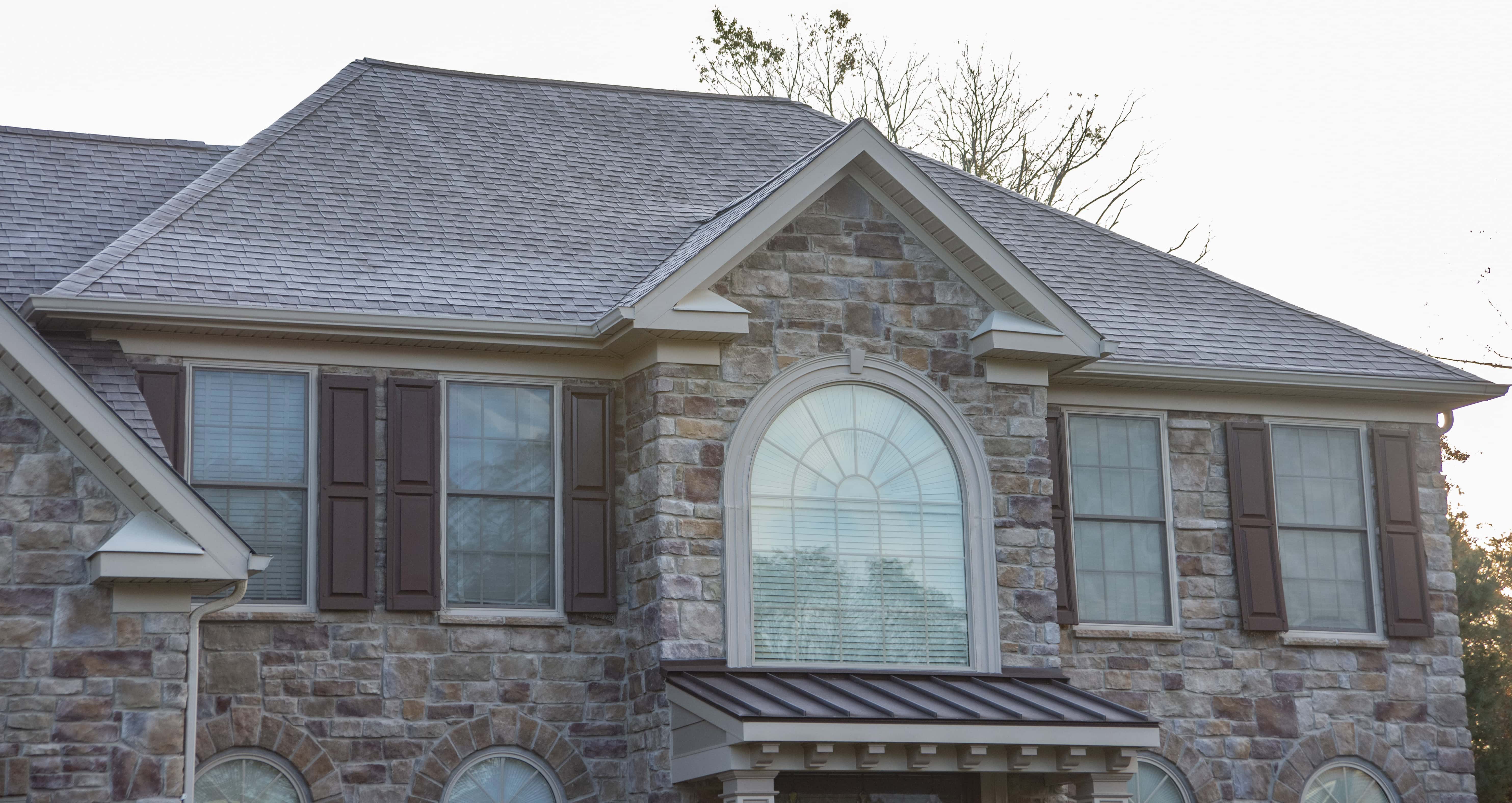 Two-story house exterior with stone facade, brown shutters, multiple windows, and a large arched window above the entrance. The roof is gray, and trees are visible in the background.
