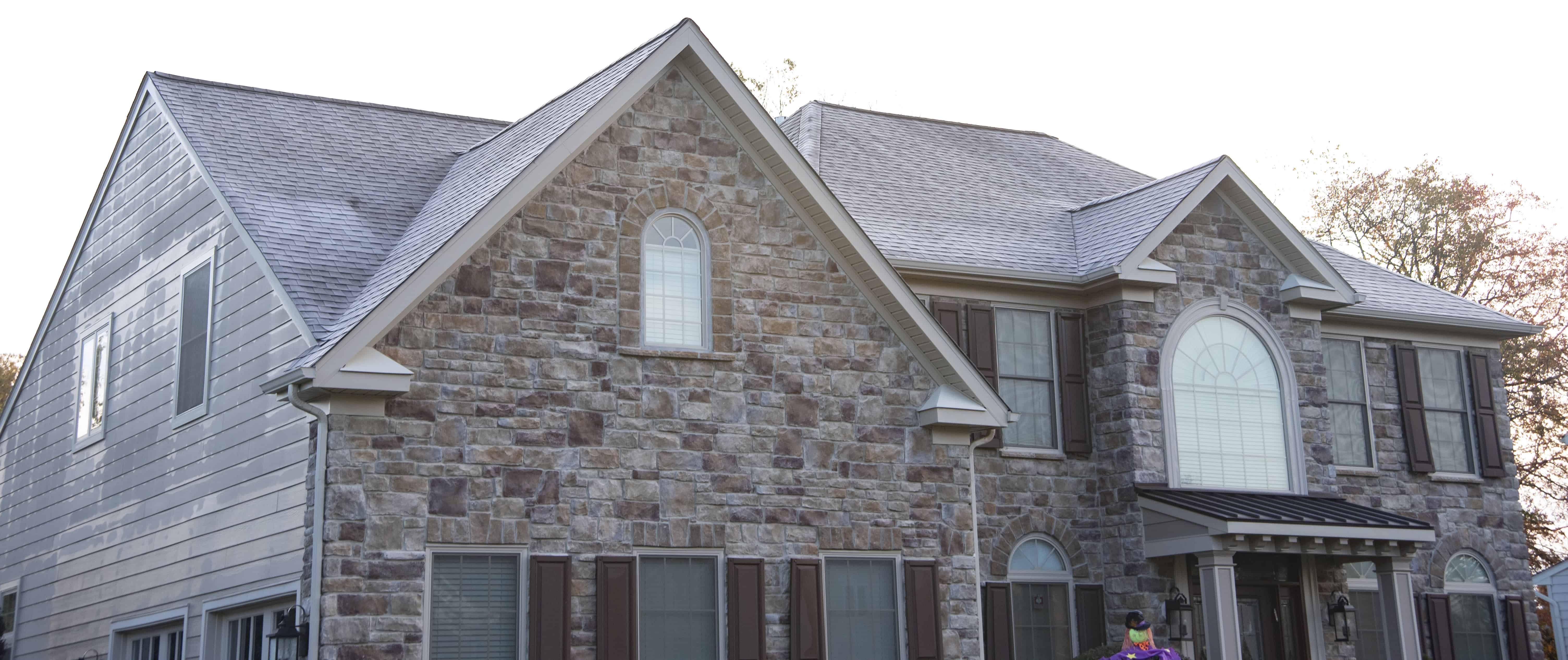 Two-story house with stone and siding exterior, multiple windows with shutters, arched windows, and a covered entryway. The roof is gray and the yard has some trees visible in the background.
