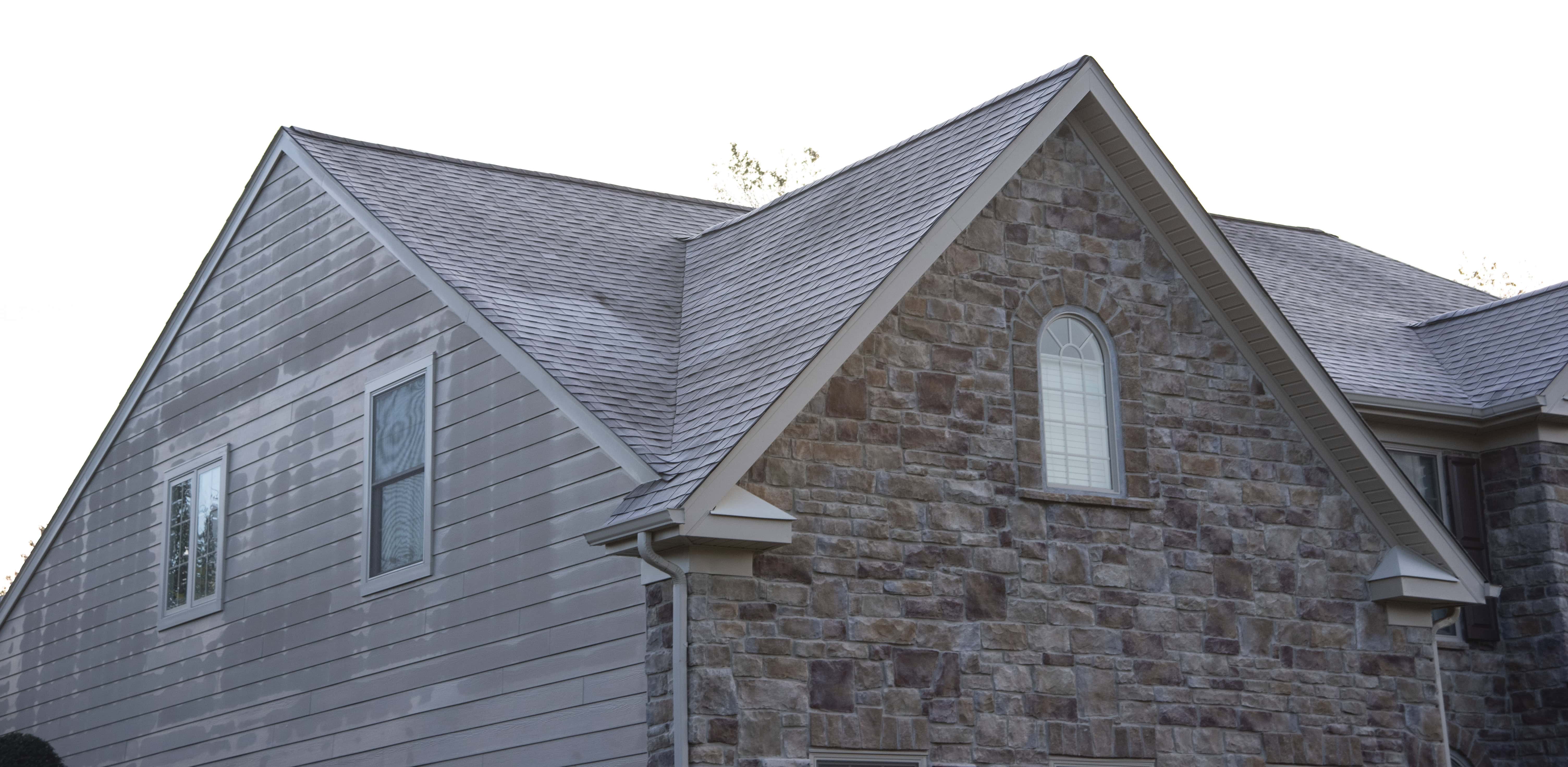 A close-up view of a house exterior featuring gray siding on one side and stone veneer on the other, with two rectangular windows and one arched window under a gabled roof.