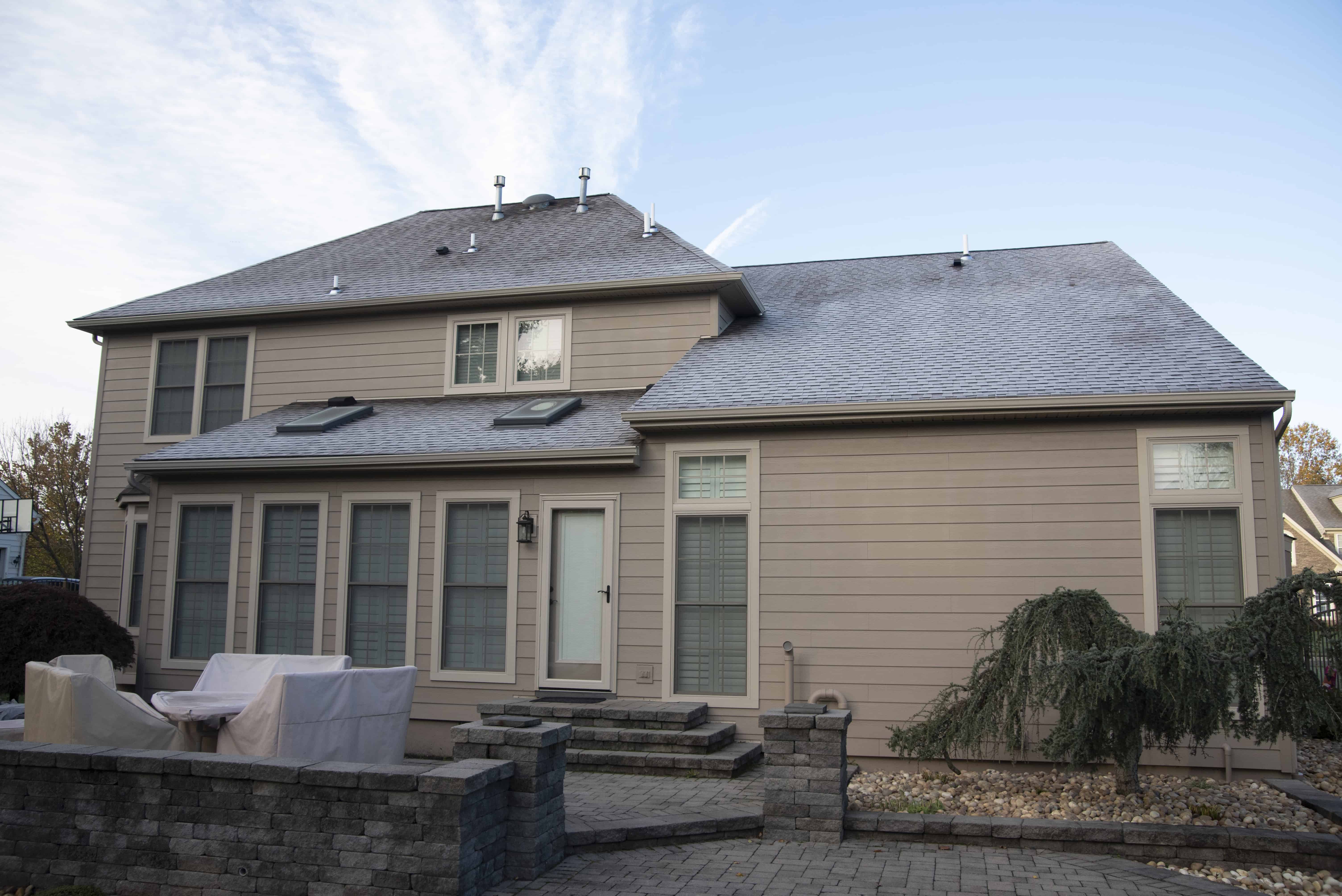 A two-story beige house with large windows and a patio area furnished with covered outdoor seating. The yard features stone pavers and a small landscaped garden with shrubs and a tree.