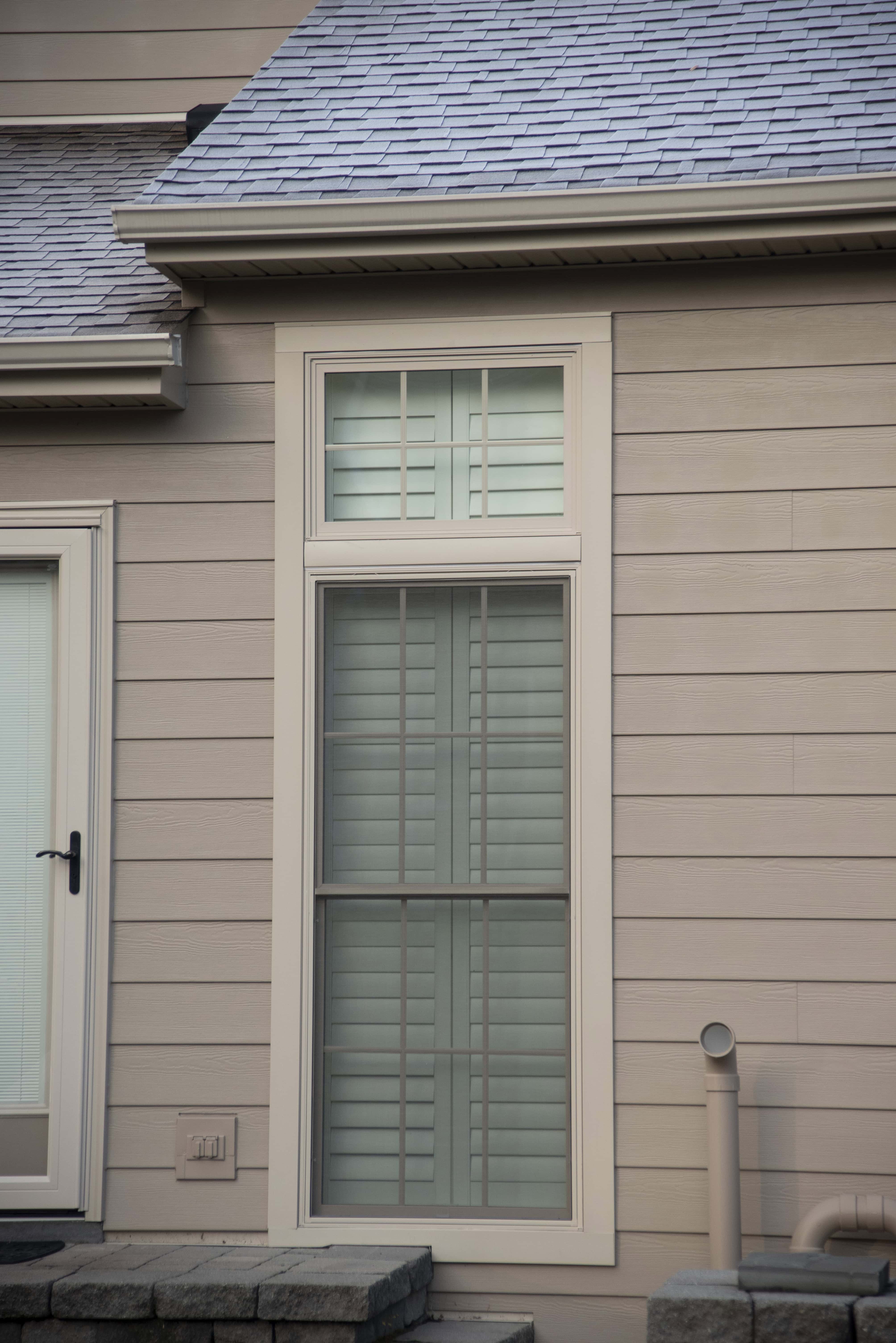 A tall, rectangular window with white trim and closed shutters is set in the beige siding of a house, next to a glass door and above a short stone wall.