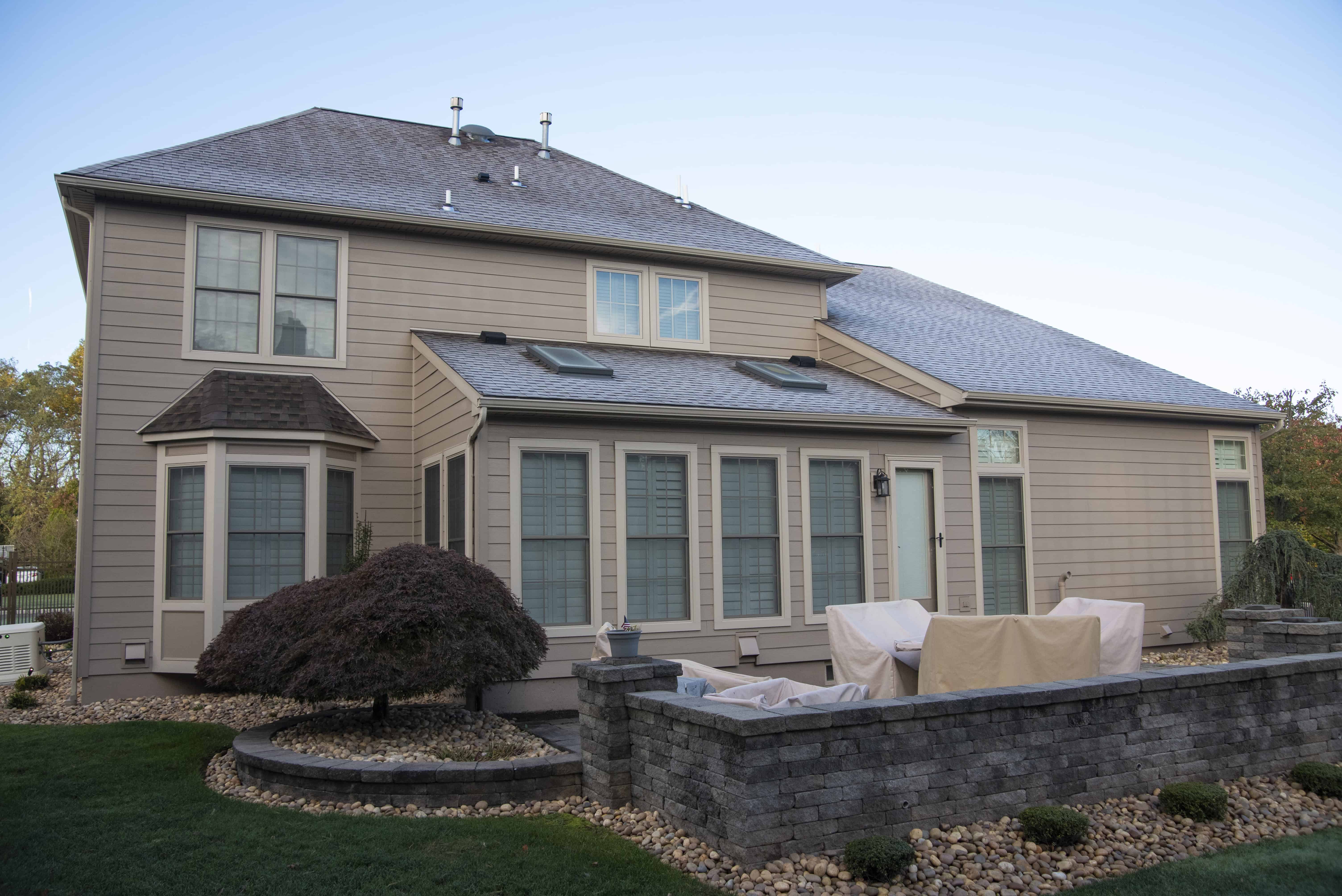 A beige two-story house with large windows and a gray roof. The backyard has a small patio with outdoor furniture covered in cloth and is surrounded by a stone wall, landscaping rocks, and trimmed bushes.