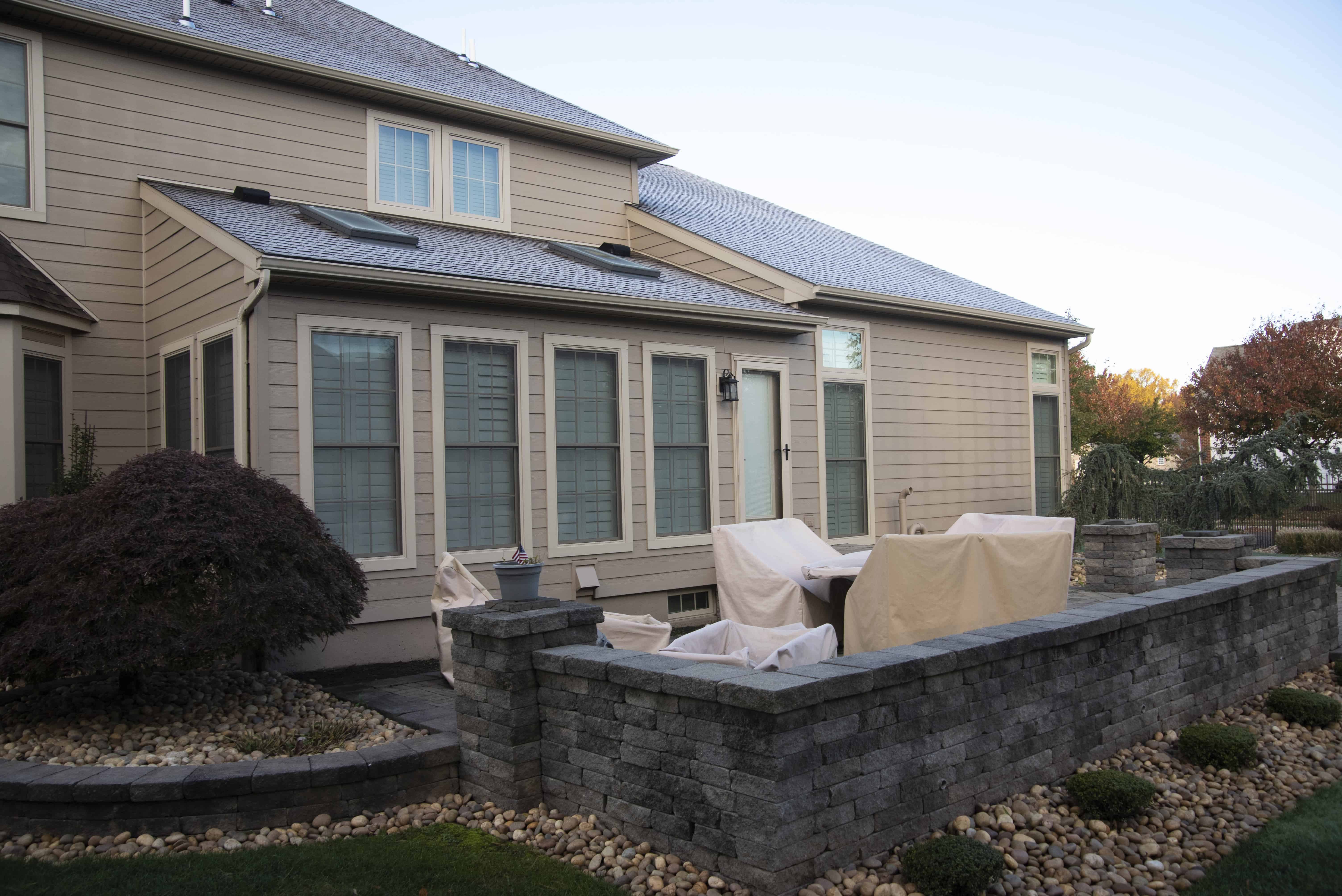 A backyard patio with covered outdoor furniture, surrounded by a short stone wall and landscaped with rocks and plants, attached to a beige house with large windows.
