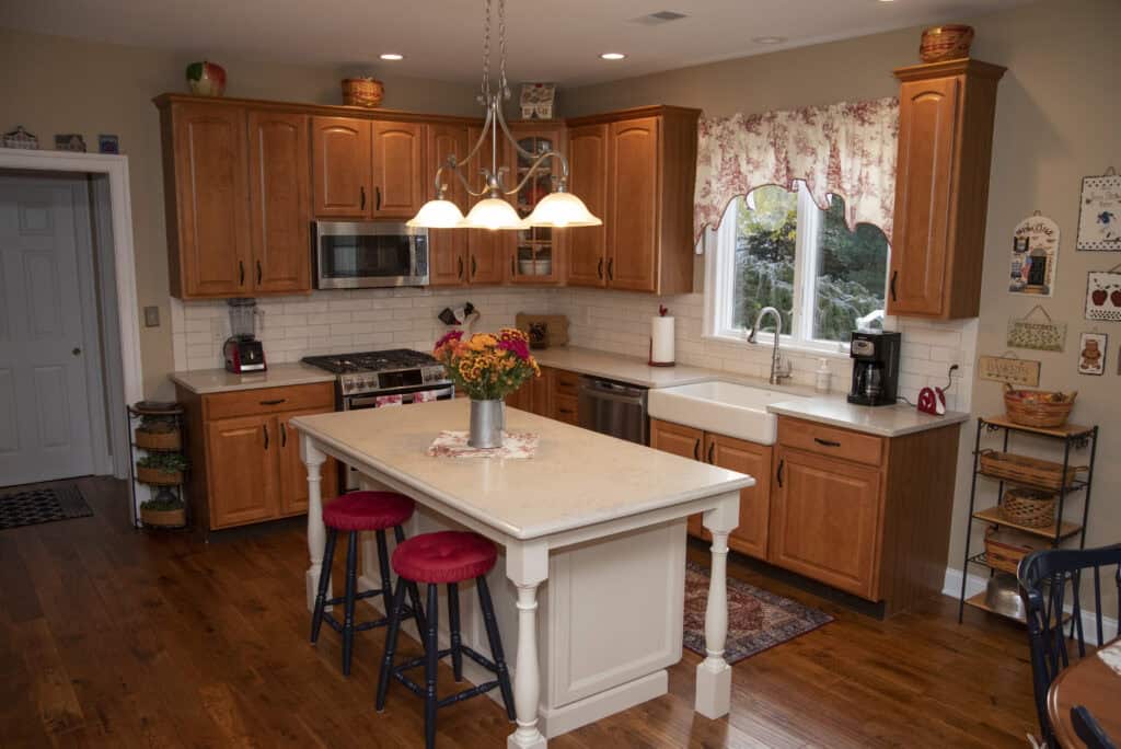 A cozy kitchen with wooden cabinets, a white island with two red stools, a vase of flowers, stainless steel appliances, farmhouse sink, coffee maker, and decorative accents on shelves and counters.