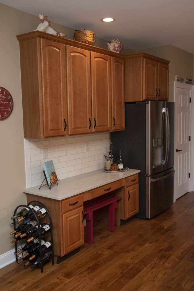 A kitchen features wooden cabinets, a stainless steel refrigerator, a small countertop with a red stool underneath, a wine rack with bottles, and white subway tile backsplash on a wood floor.