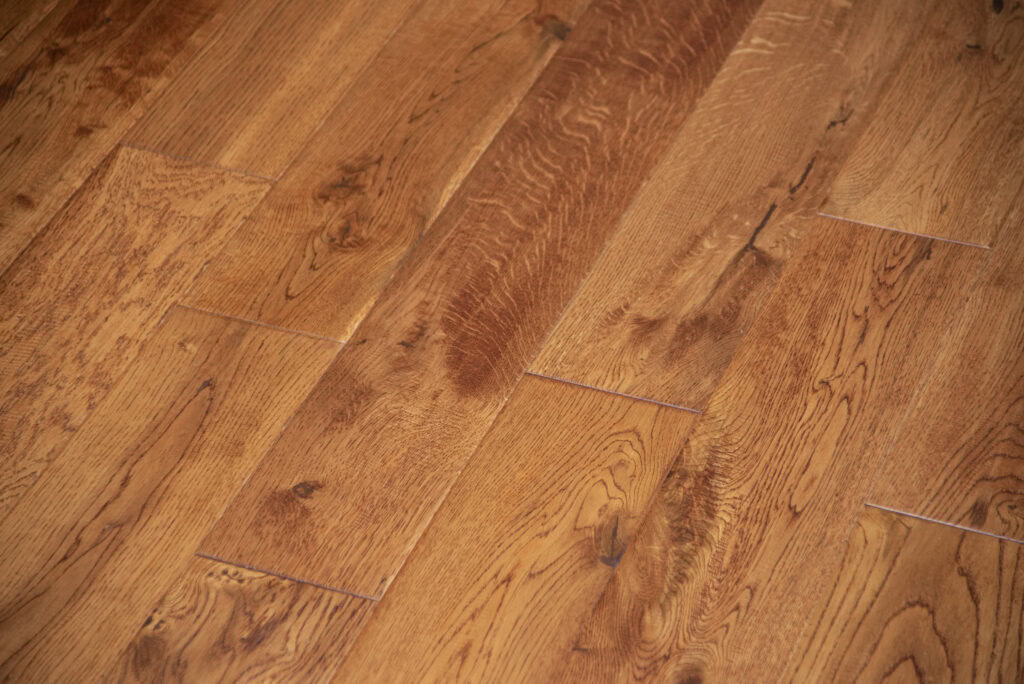 Close-up view of a light brown wooden floor with visible grain patterns and natural knots. The planks are arranged side by side, showing a smooth, polished surface and subtle variations in color.