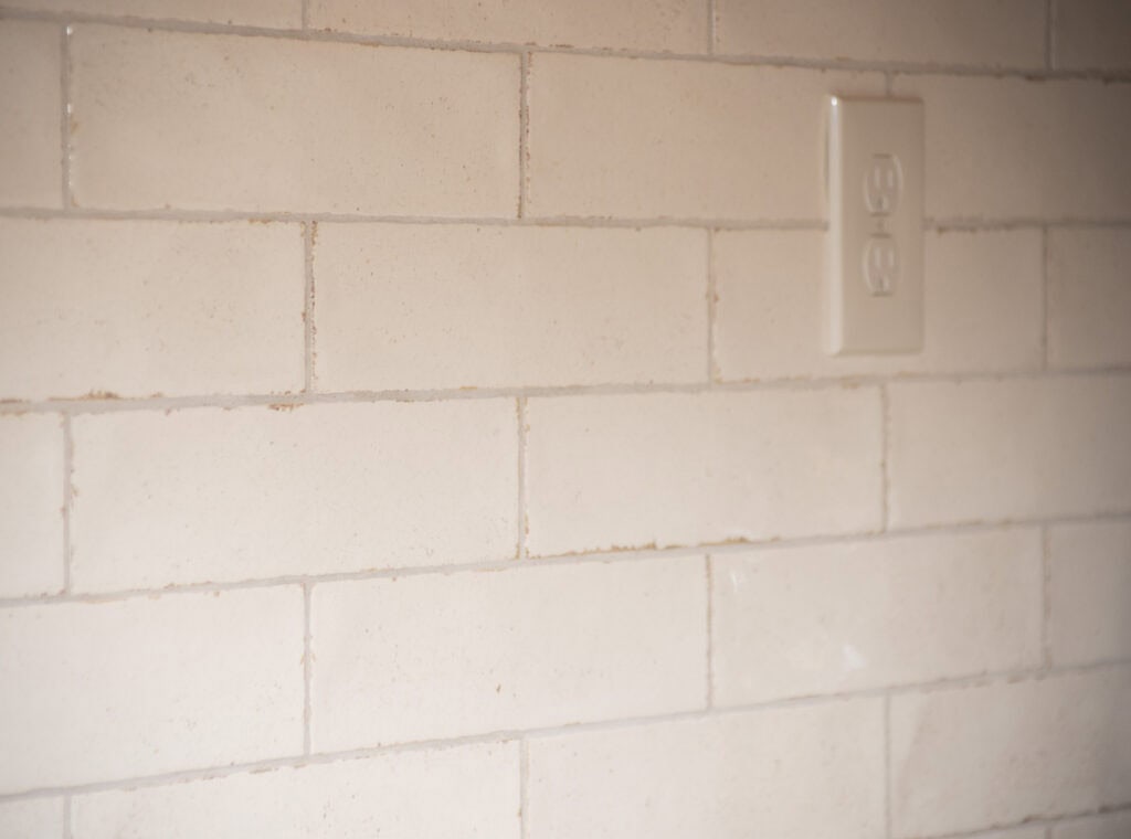 Close-up of a beige brick-patterned tiled wall with a white electrical outlet cover located in the upper right portion of the image. The grout lines between the tiles are visible.
