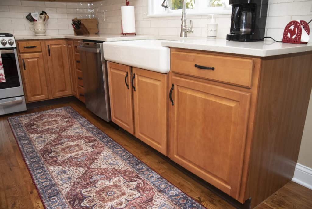 A kitchen with light wood cabinets, a farmhouse sink, a dishwasher, a stove, a coffee maker, and a patterned rug on a wooden floor. White subway tile backsplash and a window are visible above the sink.