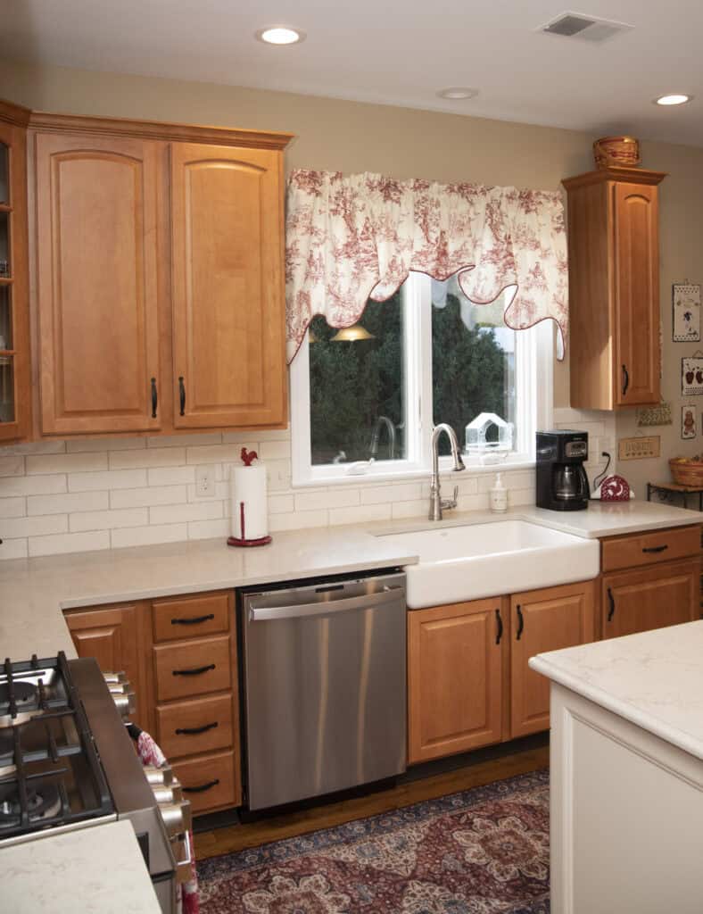 A cozy kitchen with wooden cabinets, a farmhouse sink, stainless steel dishwasher, white countertops, a coffee maker, and a window with a floral valance overlooking greenery outside.