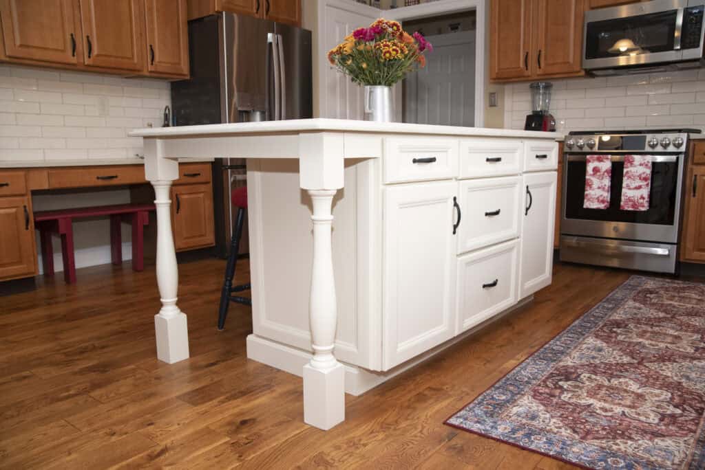 A kitchen with a white island featuring ornate legs, wooden cabinets, stainless steel appliances, a colorful rug, and a vase of flowers on the island. The floor is wood and there is a red bench in the background.