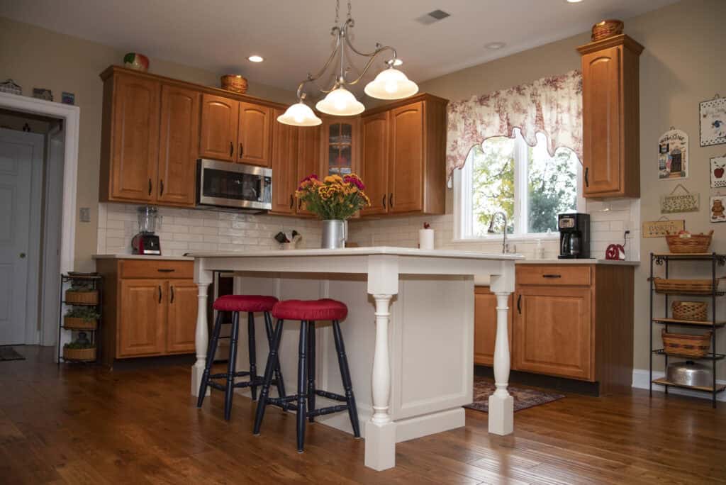 A warm, inviting kitchen with wooden cabinets, white countertops, a central island with two black stools featuring red cushions, a vase of flowers, hanging lights, and sunlight coming through a window with floral curtains.