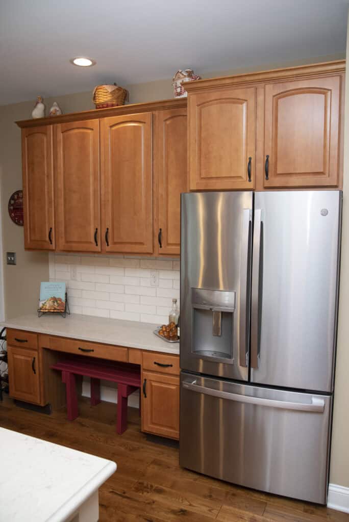A modern kitchen with light brown wooden cabinets, a stainless steel French door refrigerator, white subway tile backsplash, wooden floor, and a small red bench under the counter.