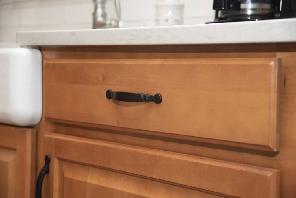 A close-up of a wooden kitchen drawer with a black handle, positioned beneath a white countertop. Part of a farmhouse sink and other kitchen items are visible in the background.