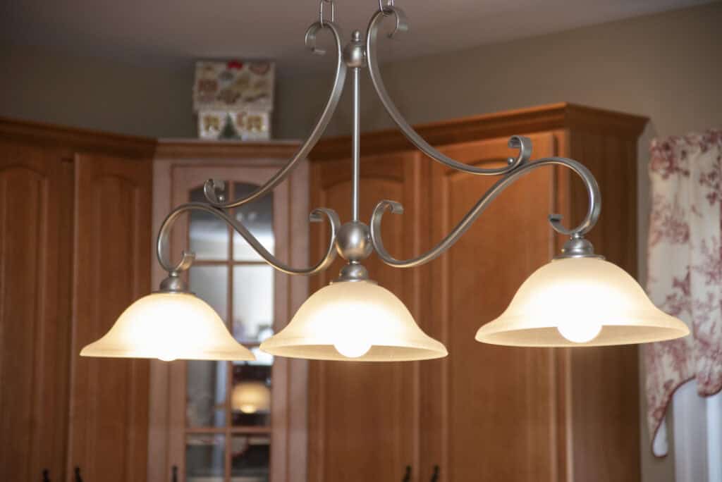 A close-up of a three-light chandelier with frosted glass shades hanging in a kitchen with wooden cabinets and a floral curtain.