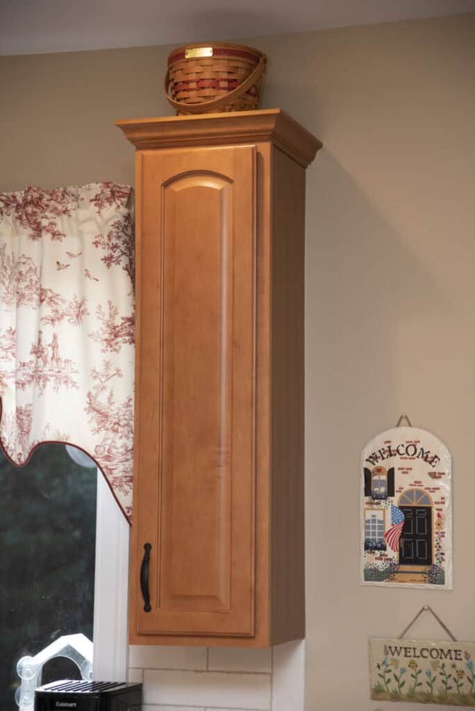 Tall wooden kitchen cabinet with a single curved door and black handle, topped with a woven basket. Nearby are two decorative welcome signs and a window with red and white patterned curtains.