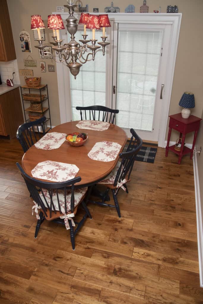 A wooden dining table with patterned placemats and five black chairs sits on a hardwood floor near glass sliding doors; a chandelier hangs above, and a red side table with a lamp is against the wall.