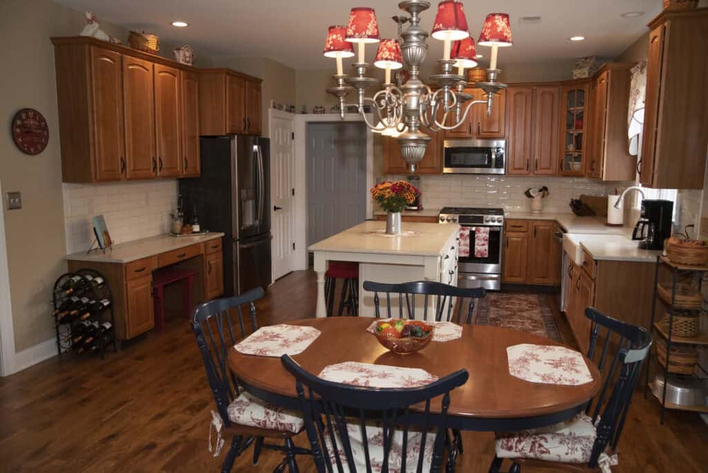 A cozy kitchen with wooden cabinets, white countertops, stainless steel appliances, and a dining table set for six. A chandelier hangs above the table, and decorative items and flowers add warmth to the space.