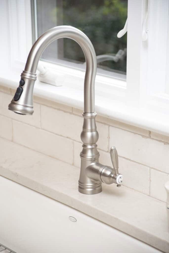 A modern brushed nickel kitchen faucet with a curved spout and a single lever handle, installed on a light-colored countertop in front of a window with cream-colored tile backsplash.