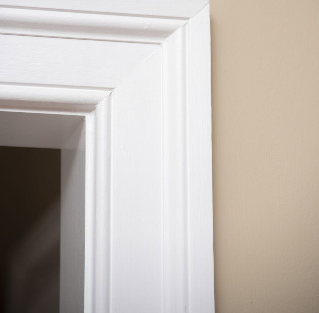 Close-up of a white door frame against a beige wall, showing the corner where the frame and wall meet. The image highlights the clean lines and smooth finish of the trim and wall.