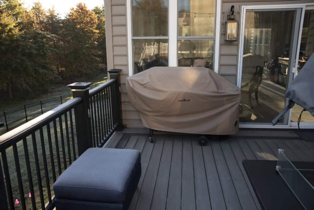 A covered grill sits on a wooden deck near a sliding glass door, with an ottoman and outdoor furniture nearby. Sunlight shines on trees in the background.