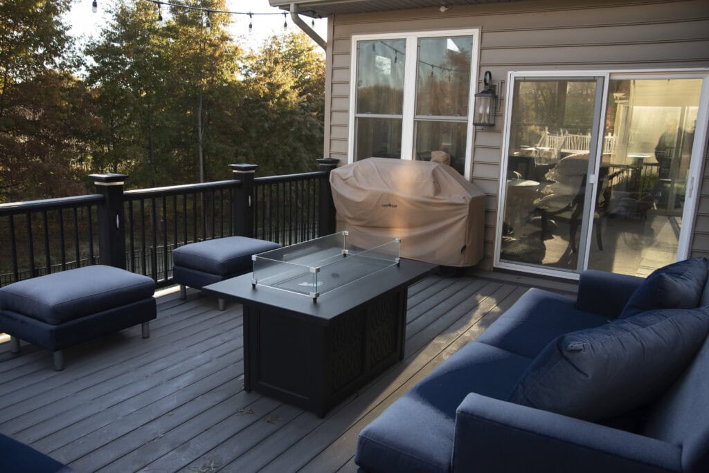 A cozy outdoor deck with navy blue cushioned seating, a glass-covered fire pit table, a covered grill, string lights, and sliding glass doors leading inside. Trees with autumn leaves surround the deck.