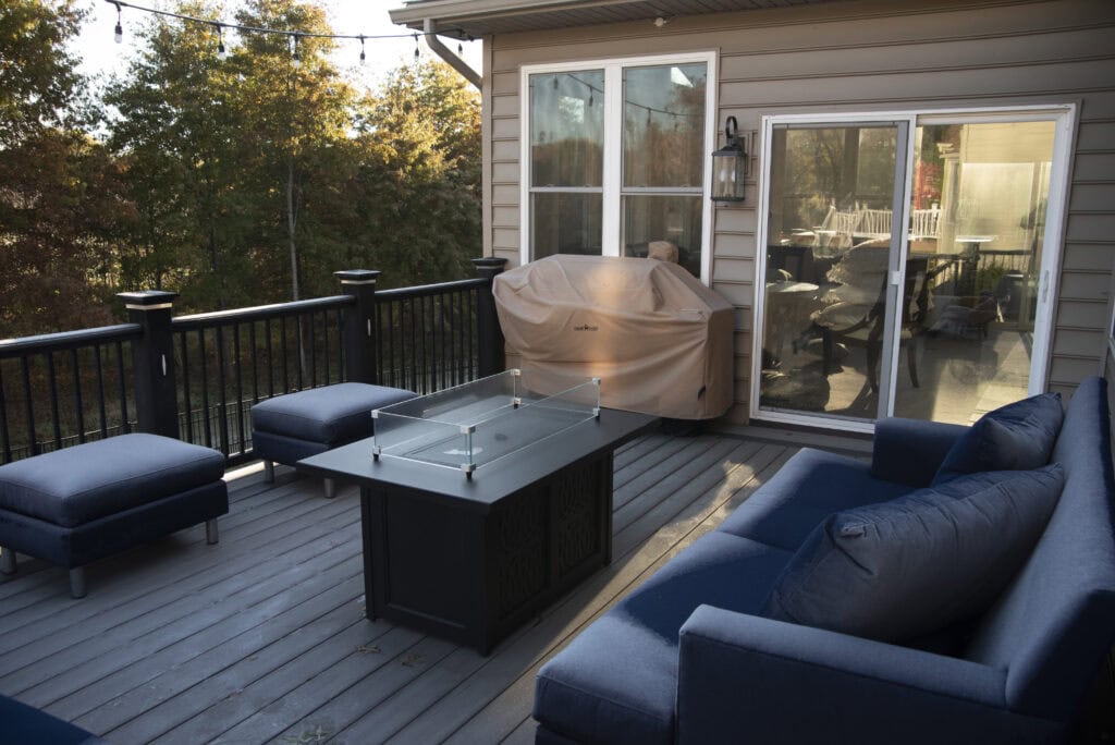 A cozy outdoor patio with a blue cushioned sofa, chairs, a glass-top fire pit table, covered grill, and sliding glass door, surrounded by trees with autumn foliage.