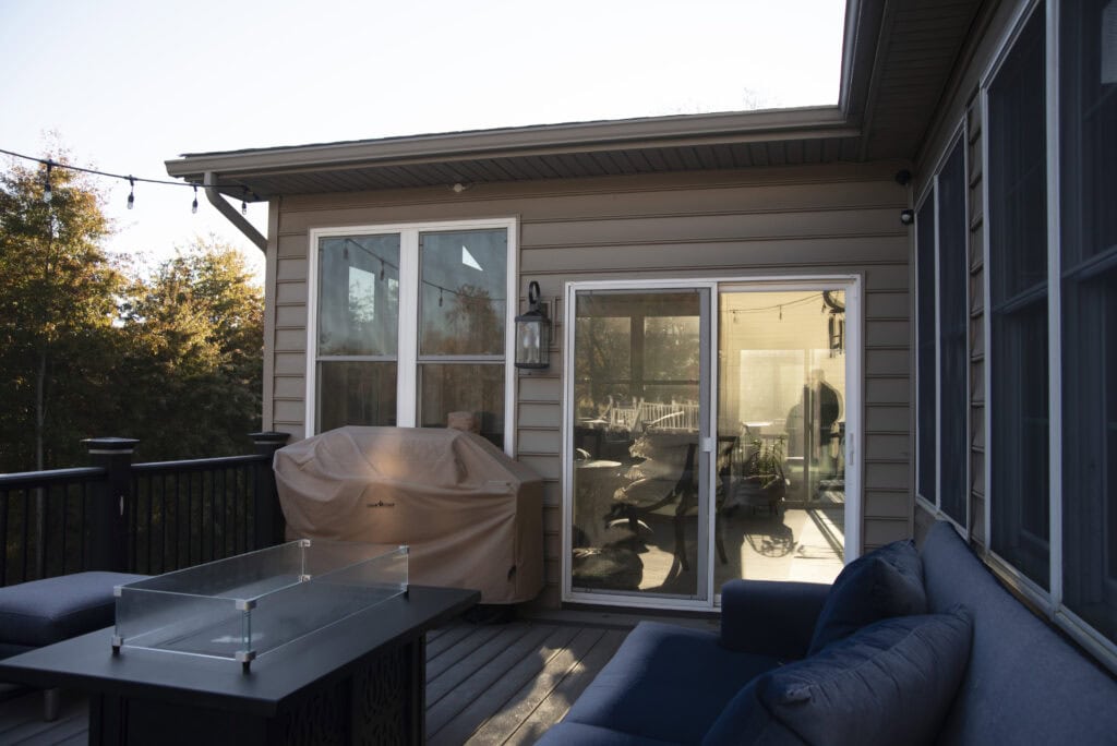A backyard deck with a covered grill, glass fire pit table, and blue outdoor sofa. Sliding glass doors and windows reflect sunlight, and a person is visible inside the house. Trees are in the background.