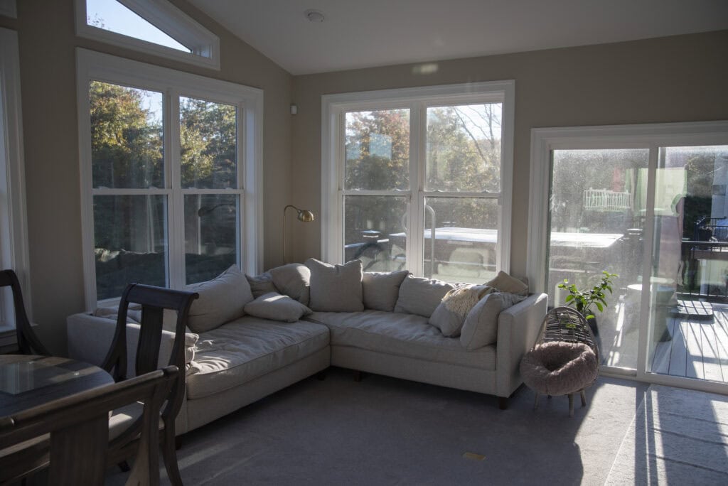 A sunlit living room with large windows and a glass door, featuring a beige sectional sofa, a small plant, and a pet bed on a light carpeted floor. Trees and outdoor furniture are visible outside.
