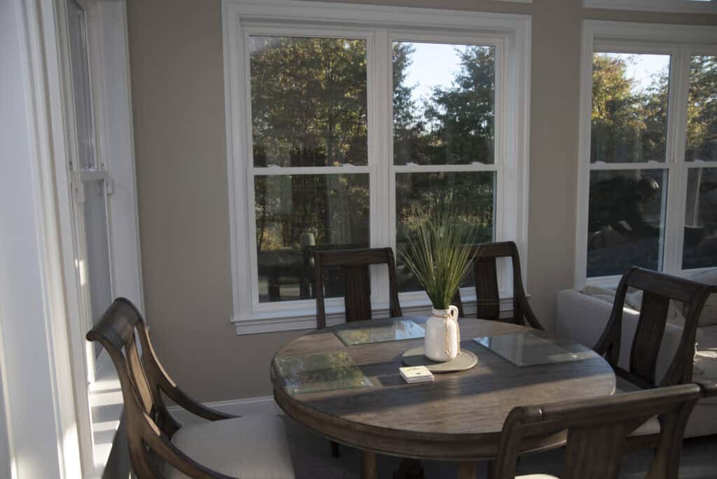 A round wooden dining table with four chairs sits by large windows, letting in natural light. A white vase with green foliage is centered on the table, and trees can be seen outside.