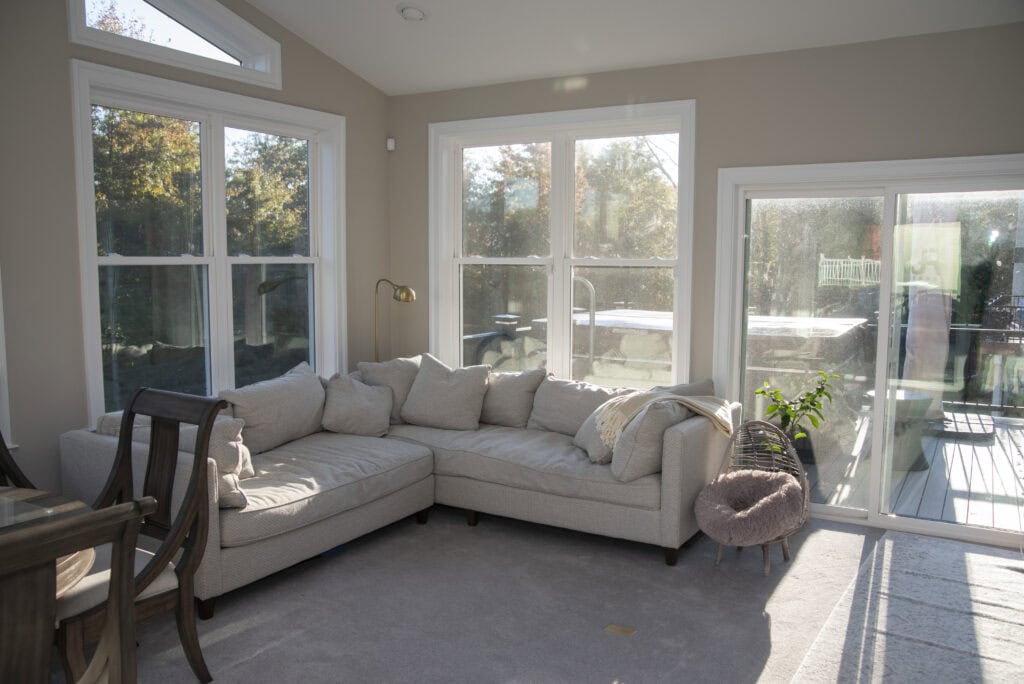 A sunlit living room with large windows, a beige sectional sofa with cushions and a blanket, a small plant, and a sliding glass door leading to a deck outside.