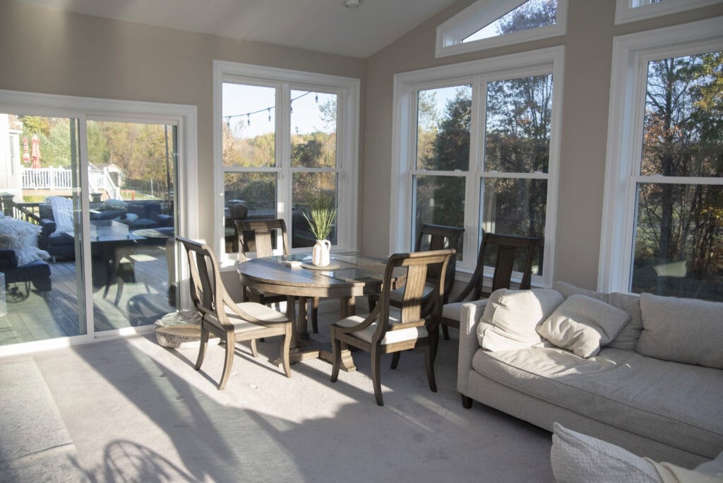 A sunlit room with large windows, a round wooden dining table surrounded by six chairs, a beige sofa, and a sliding glass door leading to an outdoor deck with patio furniture. Trees are visible outside.