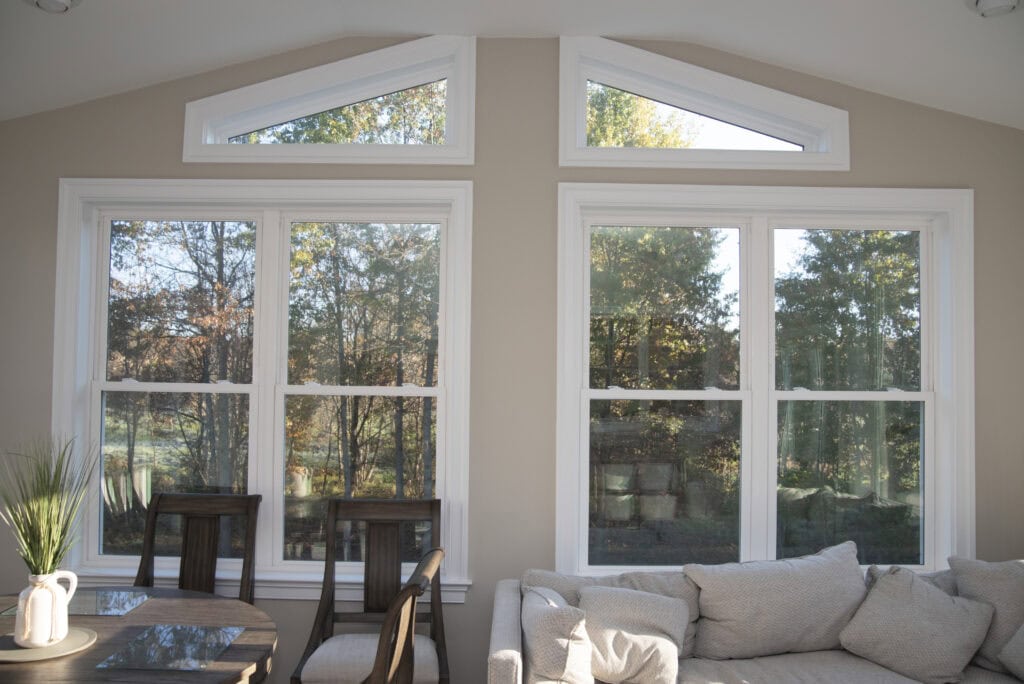 Two large rectangular windows beneath two triangular windows let sunlight into a cozy living room with a beige sofa, wooden chairs, and a small table. A view of trees and greenery is visible through the windows.