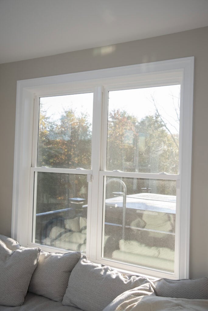Sunlight streams through a large double window above a light-colored sofa, revealing an outdoor view of a hot tub, deck railing, and autumn trees with changing leaves.