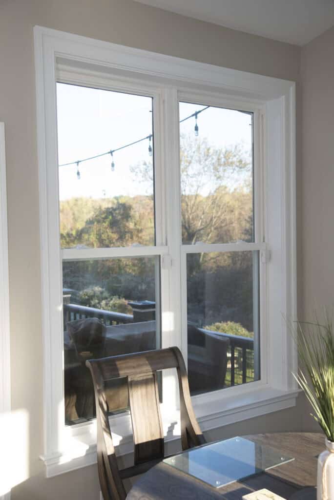 A sunlit dining area with a glass table and chairs sits beside a large window overlooking a deck and a scenic, tree-filled backyard with string lights hanging outside.