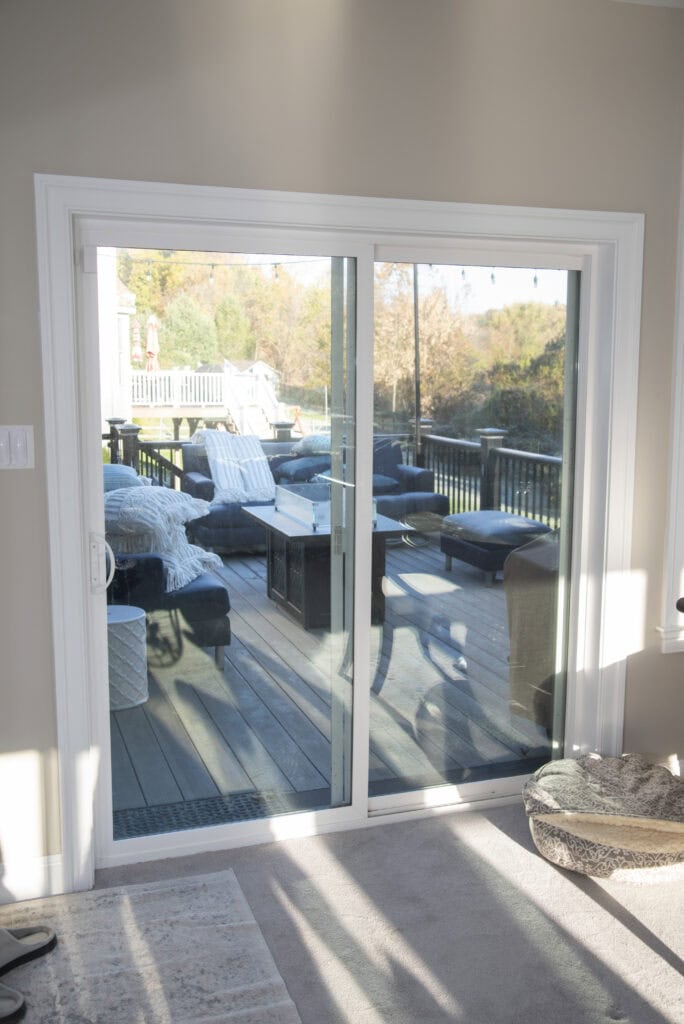 A sliding glass door opens to a sunlit wooden deck with outdoor furniture, including chairs, ottomans, and a table. Trees with autumn foliage are visible in the background.