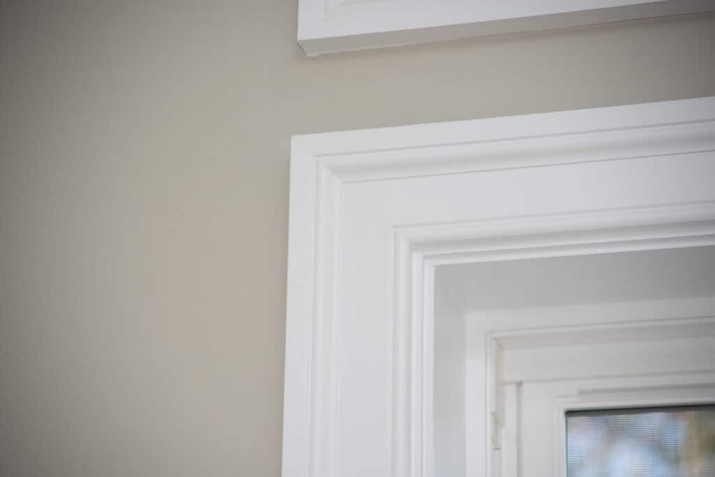 Close-up of white door trim and window frame against a beige wall, highlighting the clean lines and molding details. Natural light enters through the window, illuminating the scene.