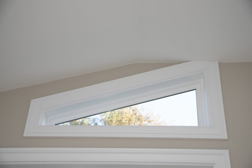 A triangular transom window with white trim is set high on a beige wall, allowing natural light to enter. Trees and blue sky are visible through the glass.