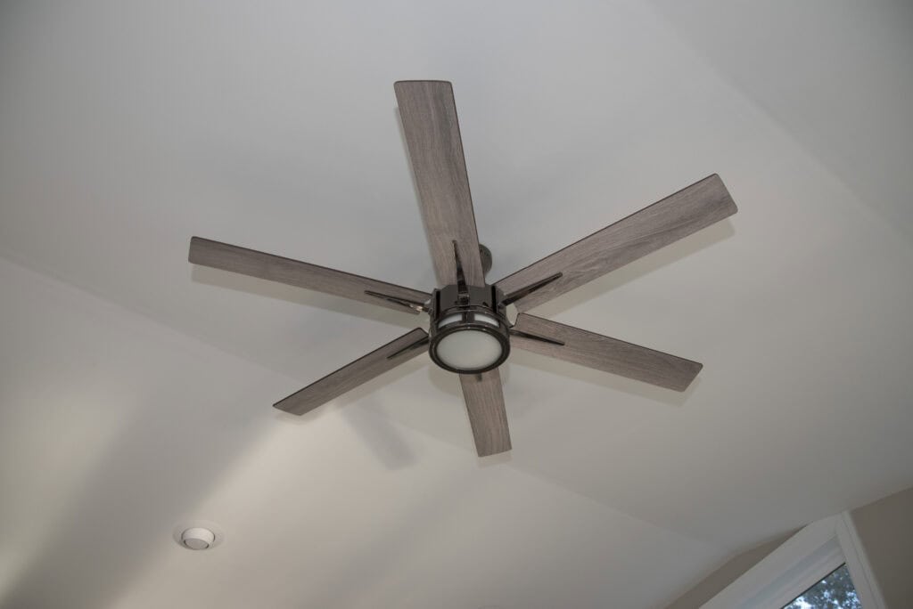 A modern, six-blade ceiling fan with a central light fixture is mounted on a white ceiling. Natural light enters the room from a window in the lower right corner.