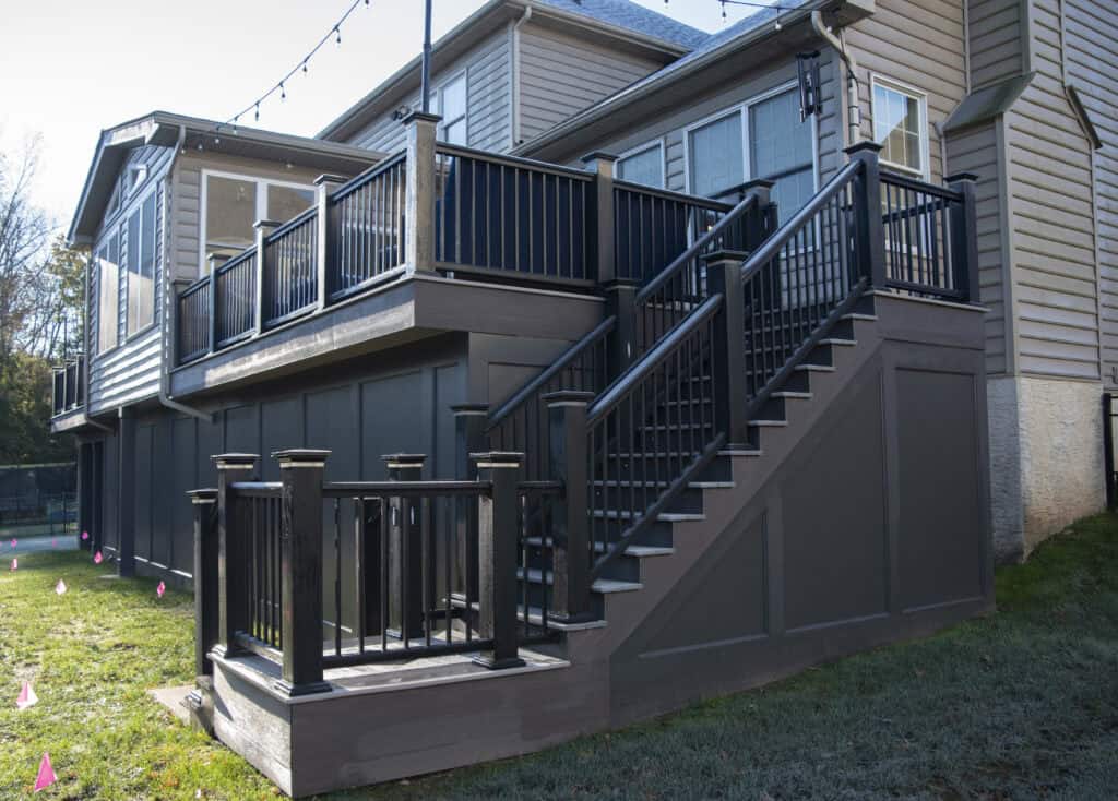 A modern, raised wooden deck with black railings and stairs attached to the back of a beige house, with string lights overhead and a grassy yard below.