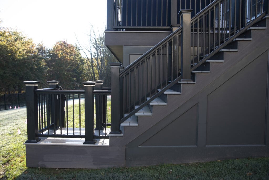 A modern outdoor staircase with black railings and dark gray steps leads up to a deck. Sunlight shines on green grass and trees in the background.