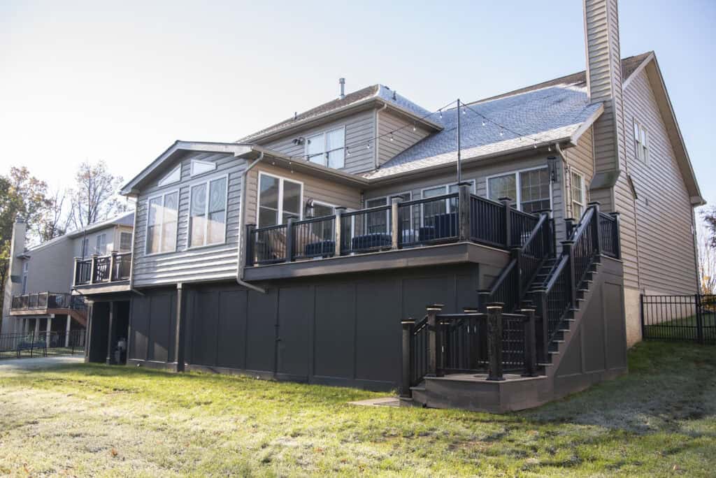 A two-story beige house with a large elevated deck, black railings, an enclosed sunroom, exterior stairs, and a fenced backyard with green grass on a sunny day.