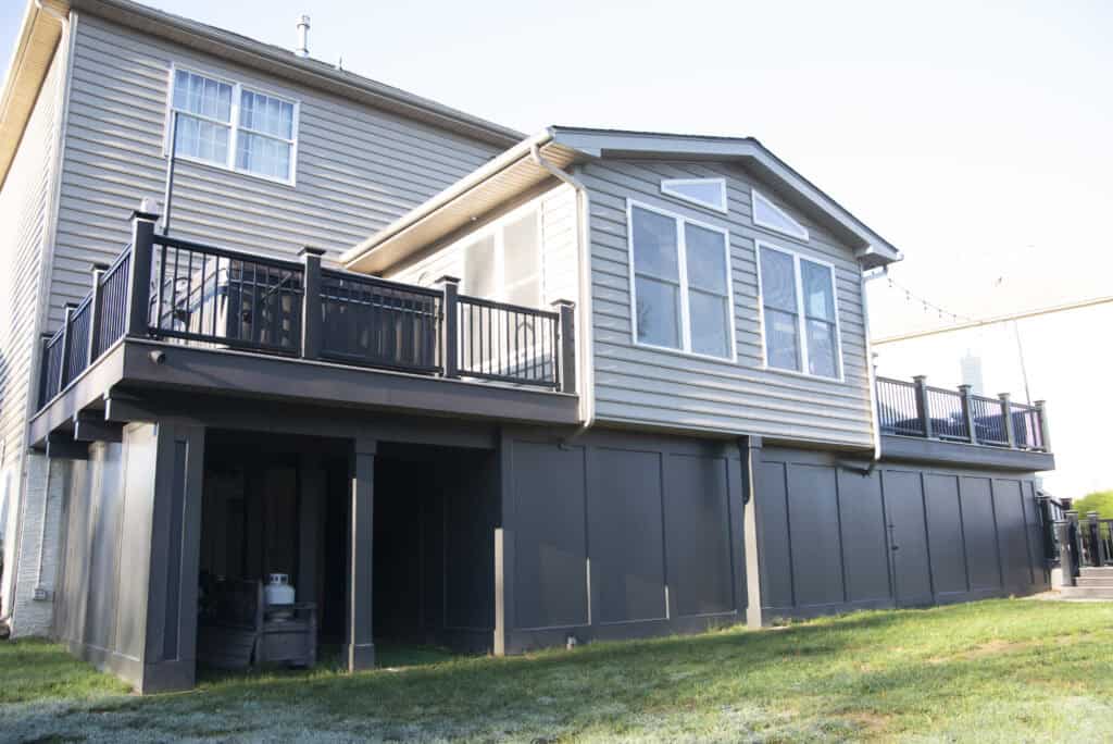A two-story tan house with a raised black deck, enclosed lower area, and a sunroom with large windows, overlooking a grassy backyard on a sunny day.