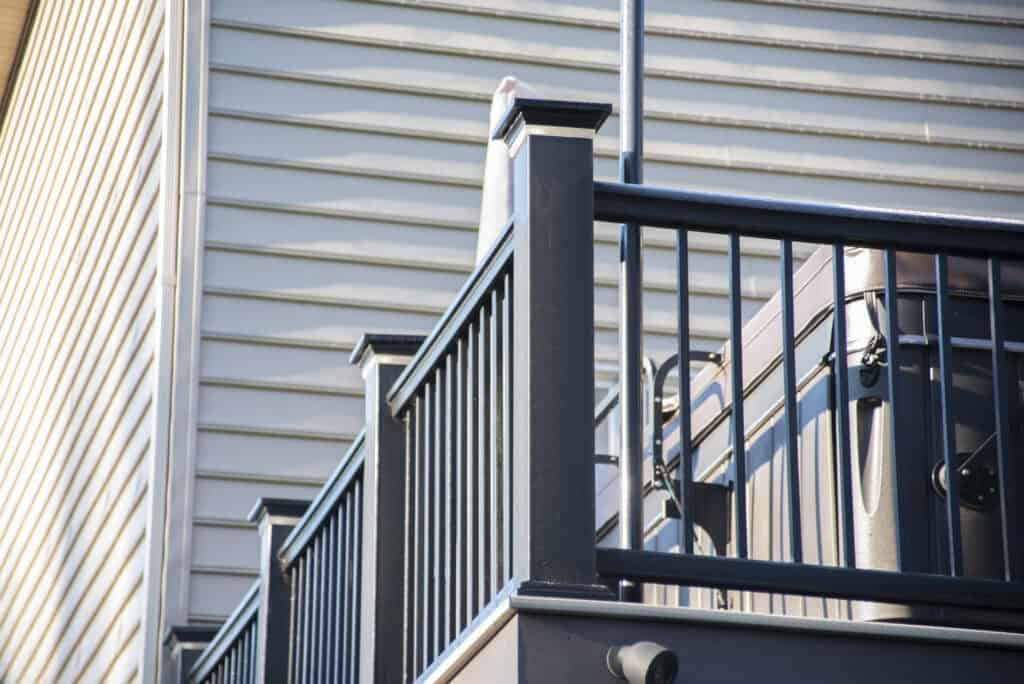 A close-up view of a modern black metal and wood deck railing attached to a house with light-colored vinyl siding. The railing surrounds an elevated deck area.