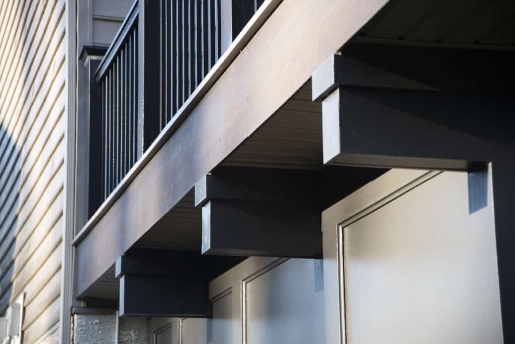 Close-up view from below of a modern building’s balcony with black railings and exposed support beams, above a row of light gray garage doors. Sunlight casts shadows on the wall and structure.