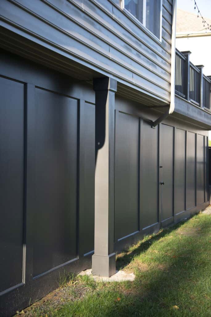 A close-up side view of a dark-colored exterior wall with decorative paneling, a supporting column, and a raised deck above, next to a grassy lawn in sunlight.