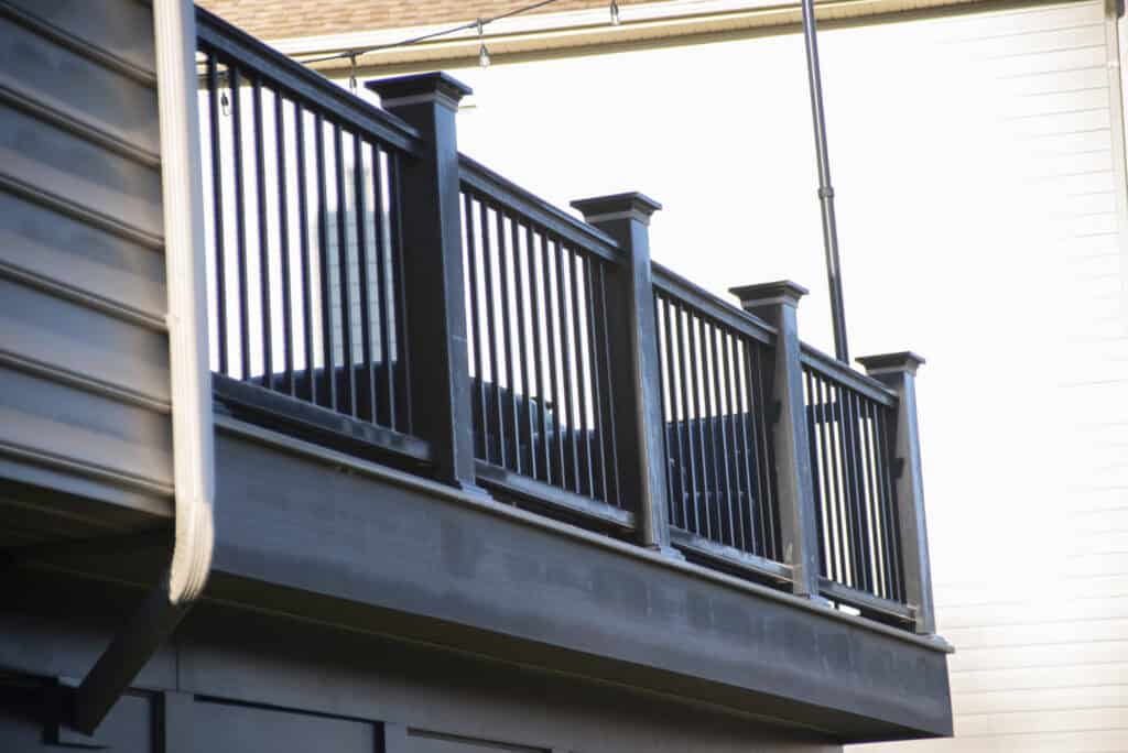 A close-up view of a modern outdoor balcony with black vertical railings and square posts, attached to a house with gray siding. The background features a white wall and part of a roof.