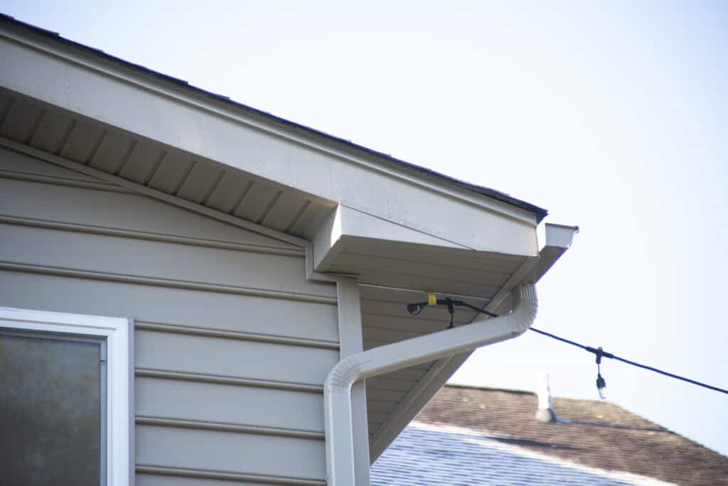 Close-up of a house exterior showing beige siding, a white gutter with a downspout, and a black cable attached near the roofline against a clear sky.