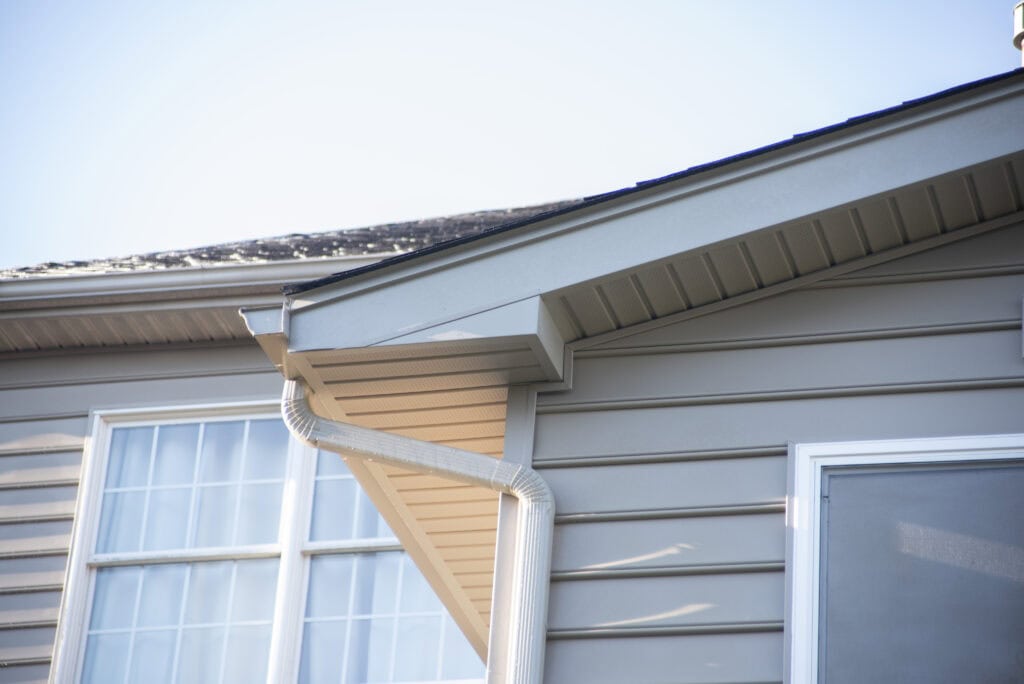 Close-up of a house exterior showing light gray siding, a sloped roof with shingles, a white gutter and downspout, and a window with multiple panes on a sunny day.